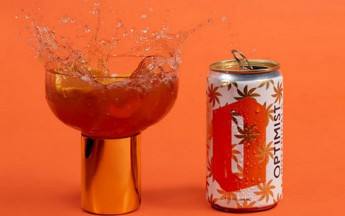A can of The Optimist's Cali Spritz beside a glass of splashing cocktail