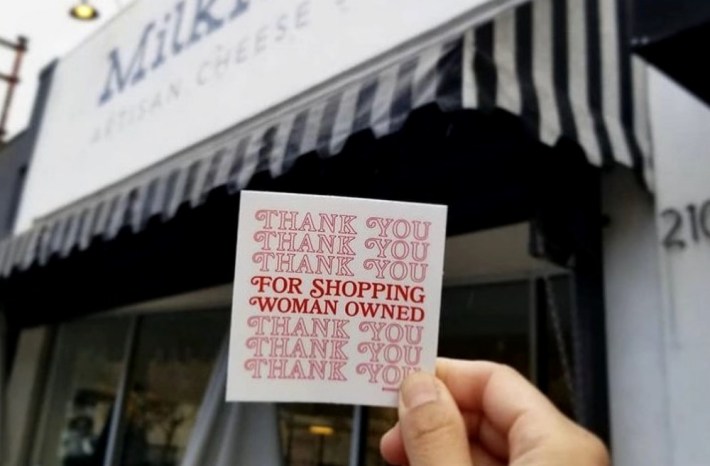 A sticker being held up in front of a striped awning that reads : Thank You For Shopping Woman Owned
