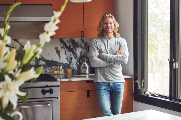 Derek Chase, a man with shoulder-length blonde hair and light beard, standing with arms folded in a kitchen