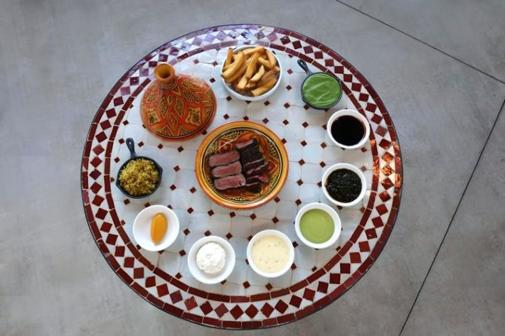 A circular plate of mezze with a tagine lid, encircled by sauces and dips, with sliced steak in the middle