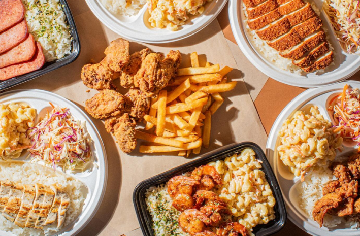 A spread of Hawaiian dishes, including fried chicken on fries, sliced fish, breaded fish, SPAM, and sides like macaroni and cold slaw