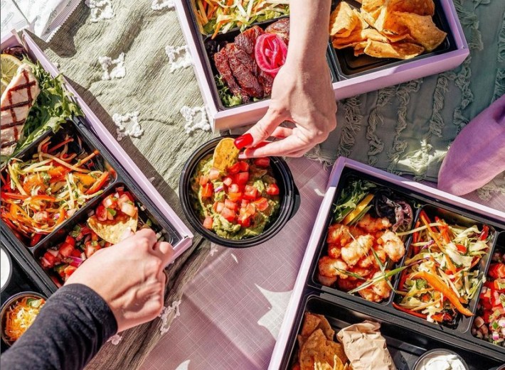 Three taco kits from Hernando's To Go with two people's hands reaching with tortilla chips