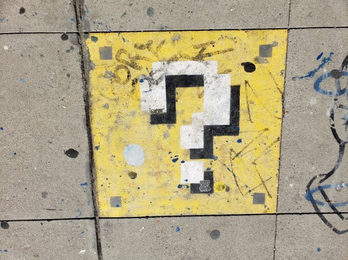 A square with a question mark inside, stenciled on the sidewalk, in the style of Super Mario Brothers