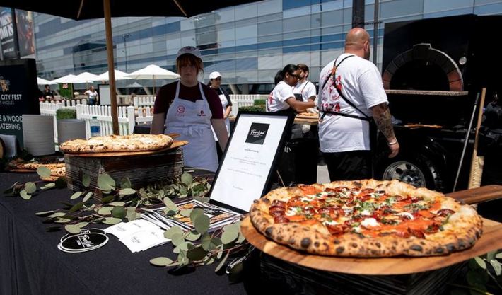 A shot of a pizza in front of chefs manning a wood-burning oven at a food festival