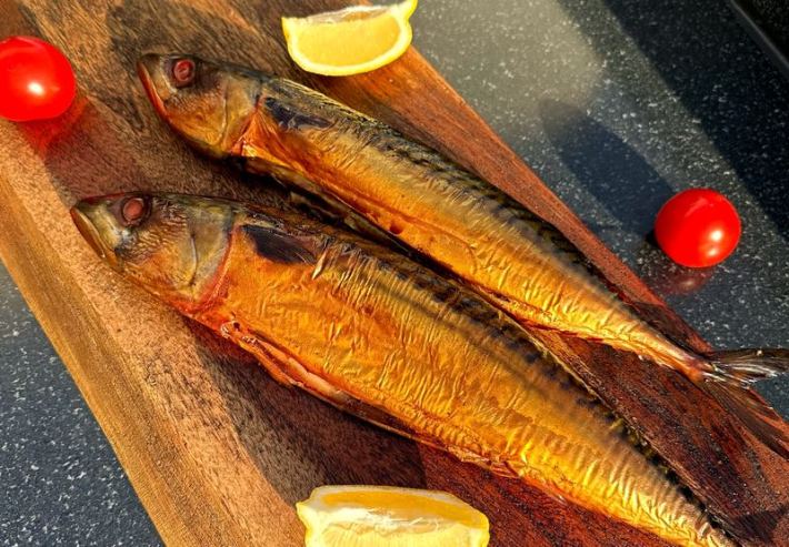 2 smoked mackerel lying side by side on a cutting board with tomato and lemons