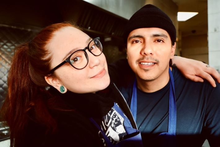 A woman with her arm over her husband's shoulder, in front of the grill in their restaurant