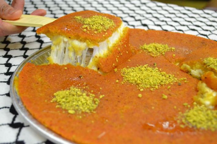A slice of knafeh being lifted from a larger knafeh, with cheese stretching from the knafeh.