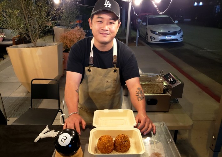 A chef in a Dodger hat stands on the street showing off two arancini he made