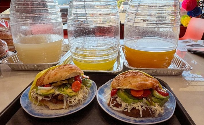 Two cemita poblanas in front of three glass jugs of agua fresca