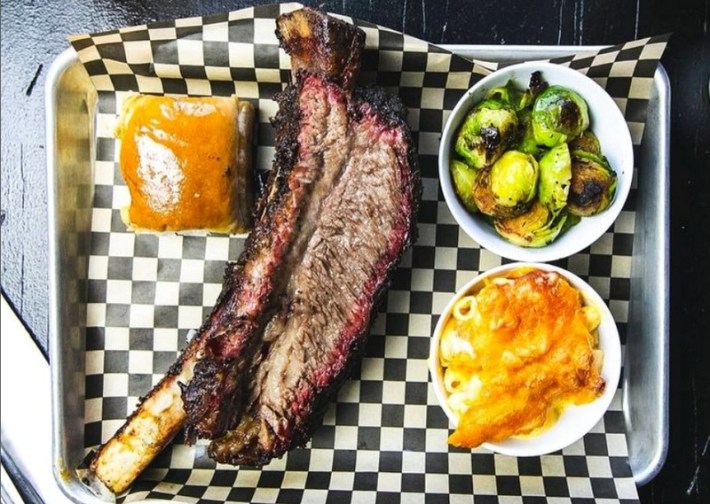 A beef rib on a tray with a bun, brussels sprouts, and mac-and-cheese on the side