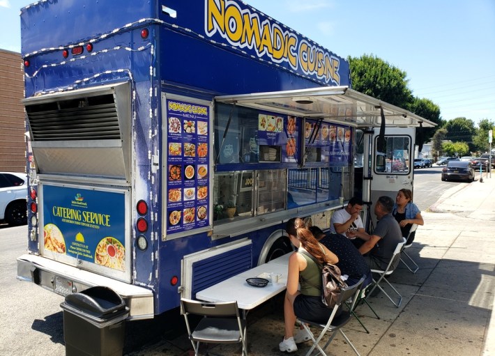 The blue Nomadic Cuisine truck, open on one side, with diners eating its food