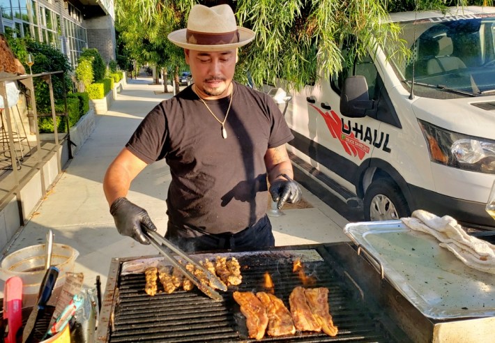 Owner Sam Oum working the grill at his Kreung Kitchen, in a panama hat with meats on the grill