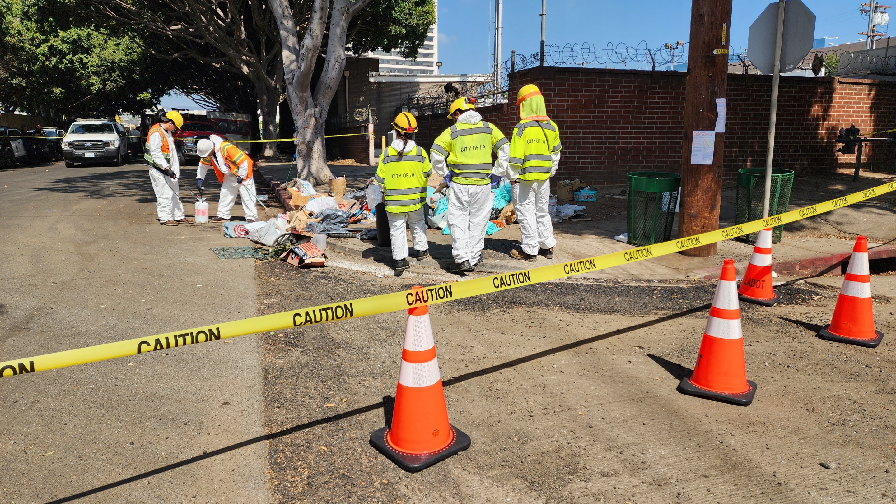 A line of caution tape separates the photographer from three sanitation workers standing in front of a pile of discarded trash and other belongings that were abandoned.