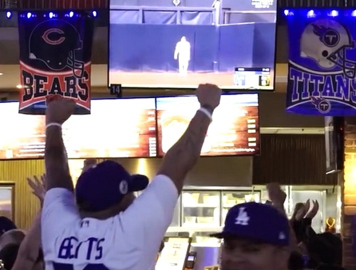 A man in a Dodger jersey raising his fists in victory while watching the Dodgers game