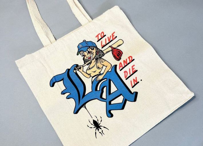 A tote bag that says "To Live And Die In LA" at Delinquent Bros. featuring a cartoon woman in clown makeup holding a baseball bat