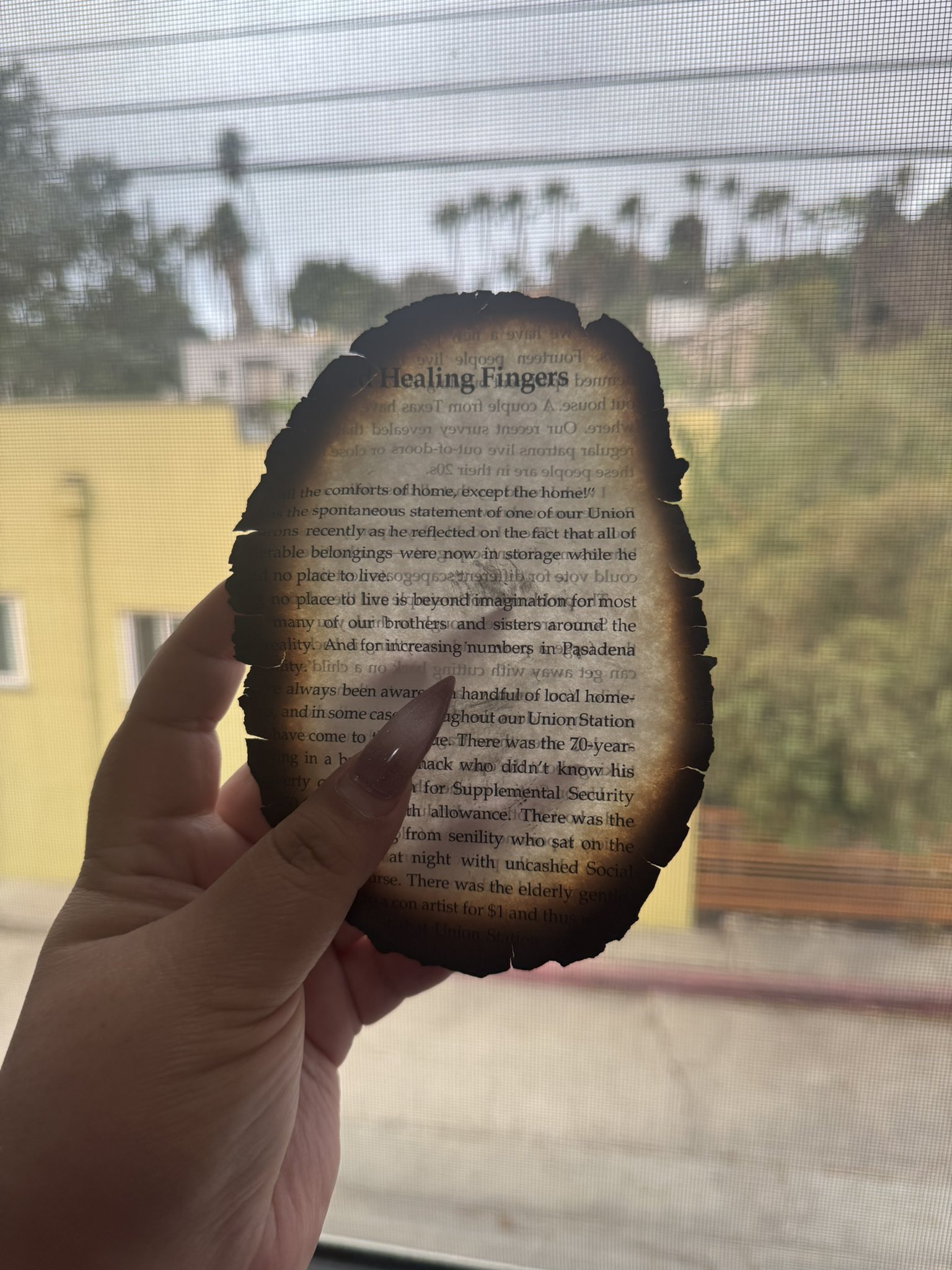 Los Angeles poet Yesika Salgado holds a partially burnt page from a book in her left hand in the foreground. In the background a yellow building can be seen through a window screen.