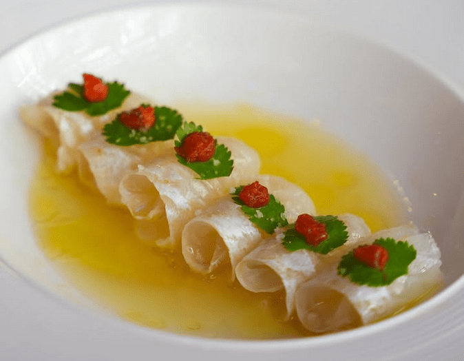 A white bowl with curls of halibut carpaccio topped with shiso and chile paste lead in a pool of yellow liquid