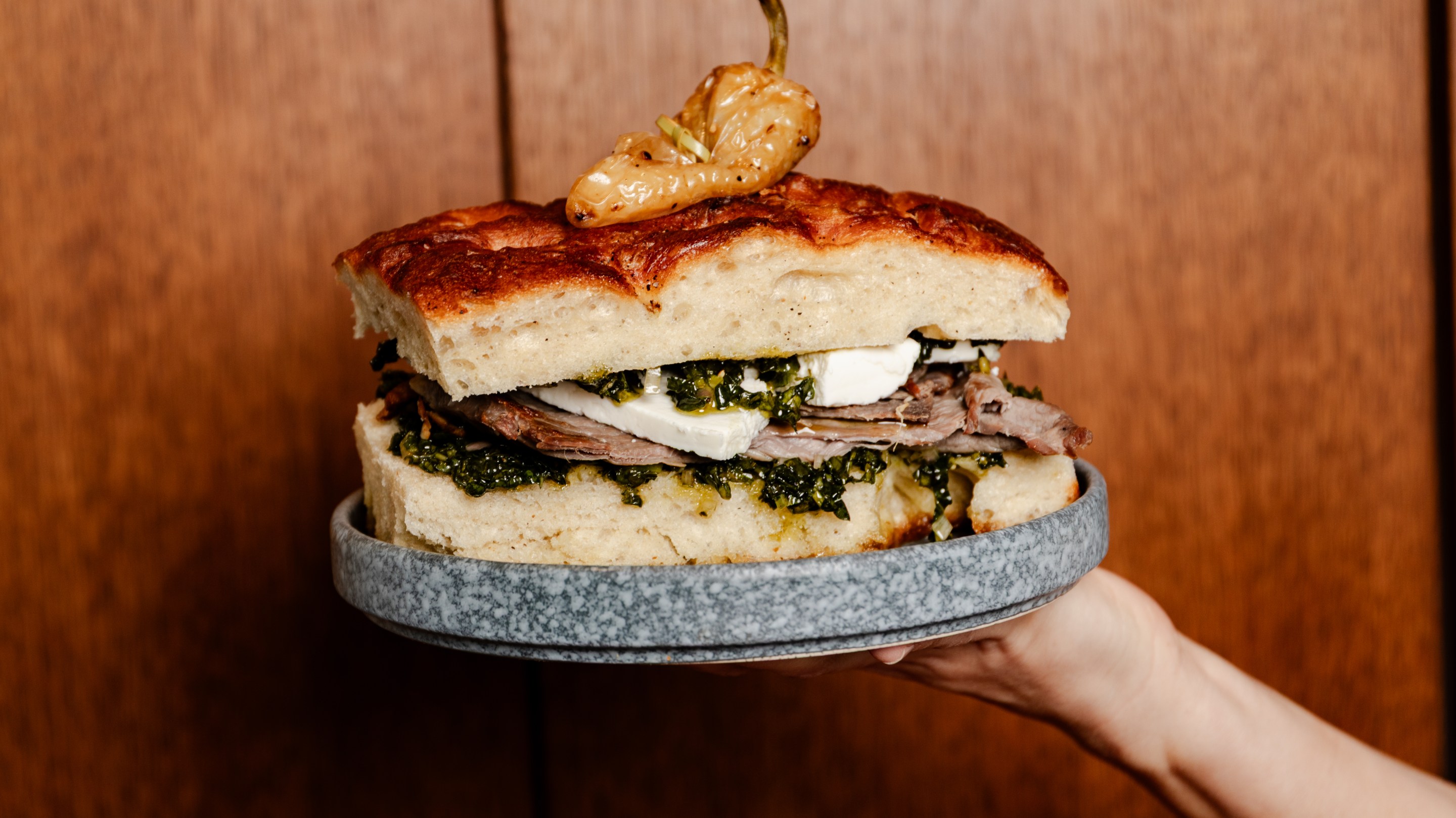 A hand holding a plate with a cecina and kale focaccia sandwich, topped with a whole chile güero, at Santa Canela.