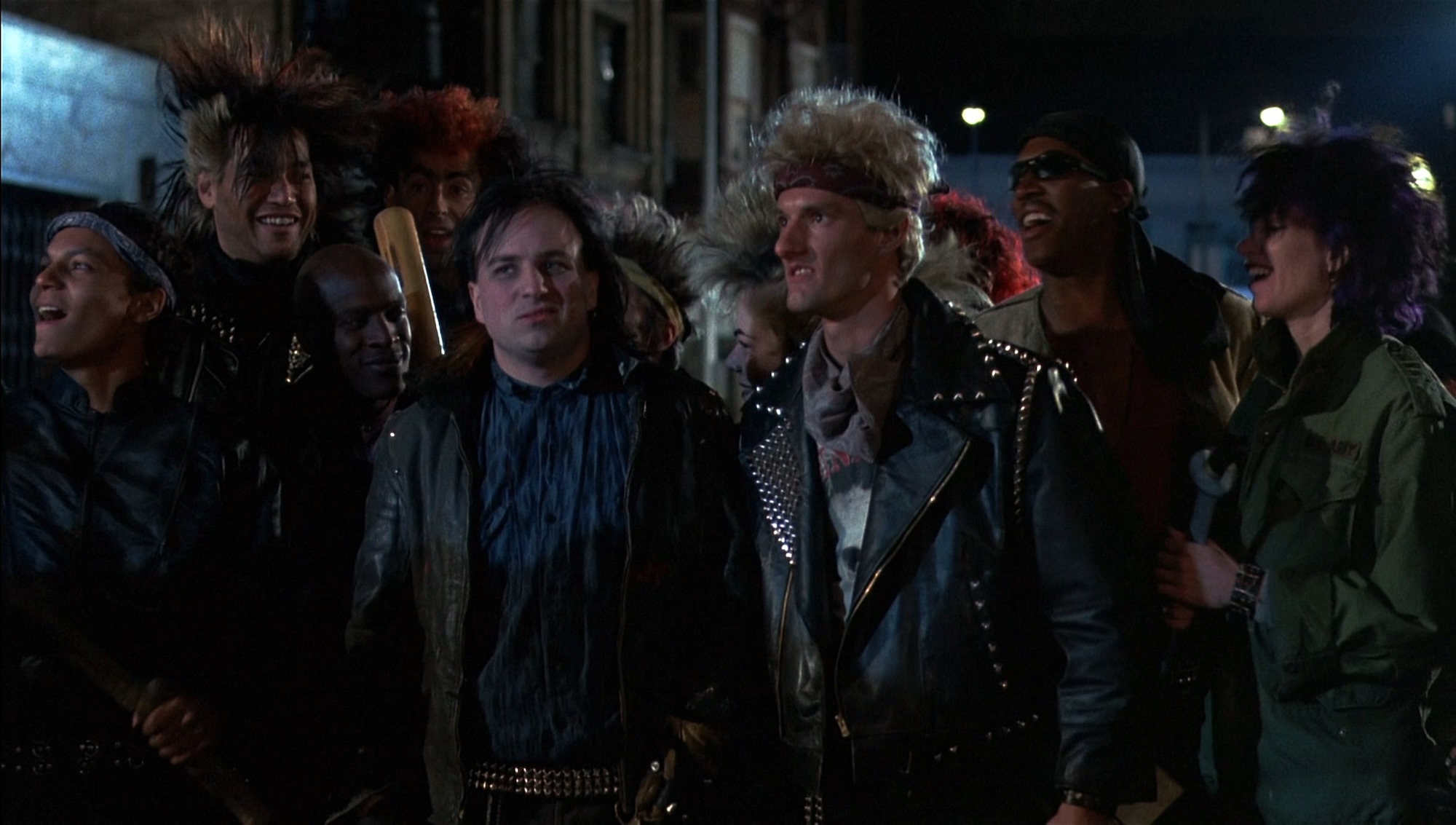 In a scene from Police Academy 2, Bobcat Goldthwait, left of center, as the Scullians’ leader, Zed, standing around other actors portraying his gang. Screenshot via Warner Bros.