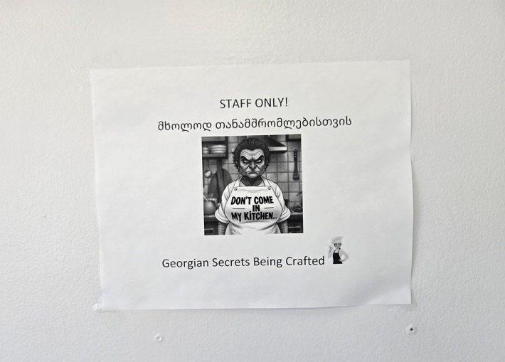 A sign on the kitchen door at Saqartvelo Georgian Cuisine in Van Nuys, showing an illustration of an angry-looking chef with an apron that says Don't Come in My Kitchen. Below it is written: "Georgian Secrets being Crafted."