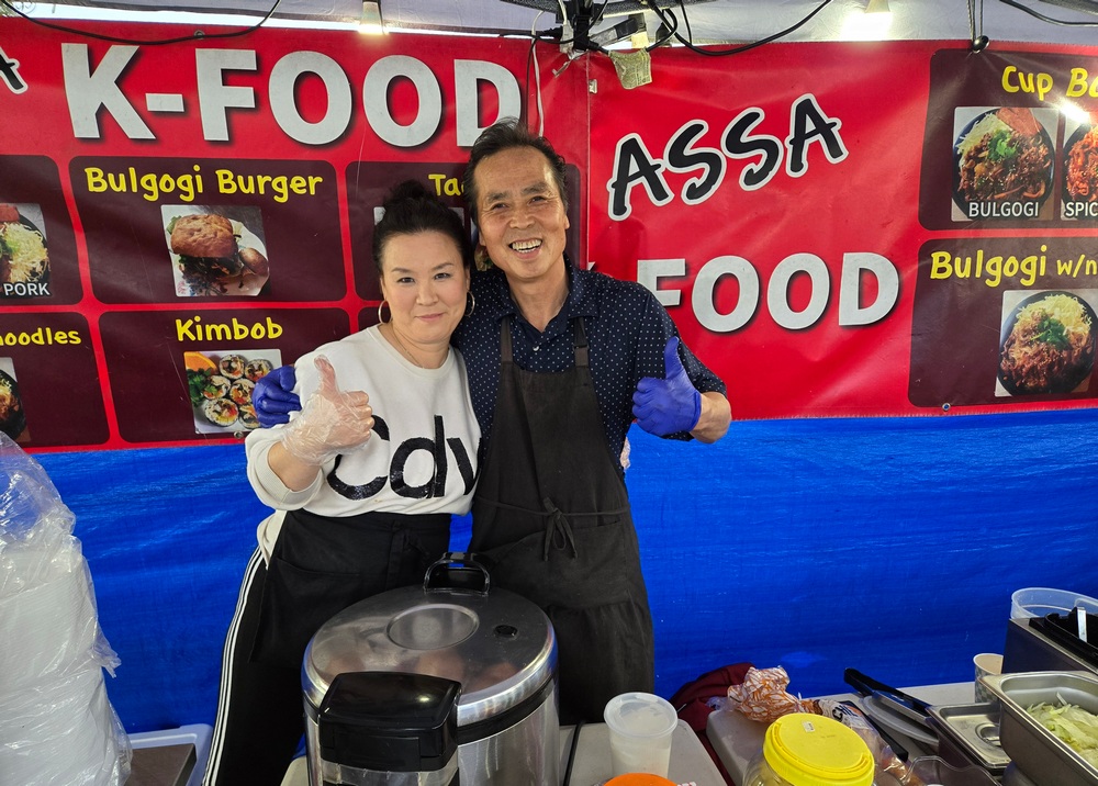 A married couple, Savannah and Ken Lee at their Korean food stand, Assa, in Venice. Both give a thumbs up in gloves, in front of their red sign.