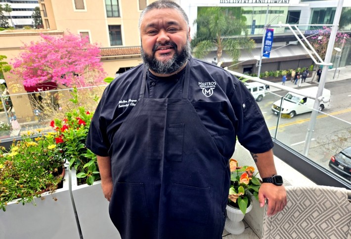 Chef Walter Mayen on the new outdoor garden patio at Mastro's Steakhouse, overlooking Canon Drive, where a jacaranda tree is in full bloom and a white truck is going by.