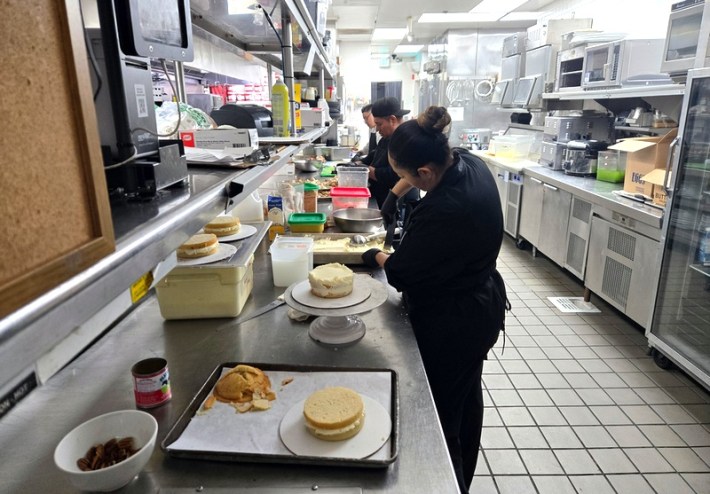 A pastry chef sticking a knife into a tray next to an assortment of cakes and pastries, with two more chefs behind her, in the kitchen at Mastros'.