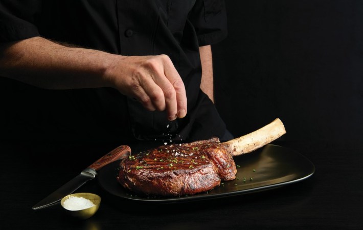 A bone-in tomahawk steak being salted at Mastro's Steakhouse, grains of salt being sprinkled on to it by a hand.
