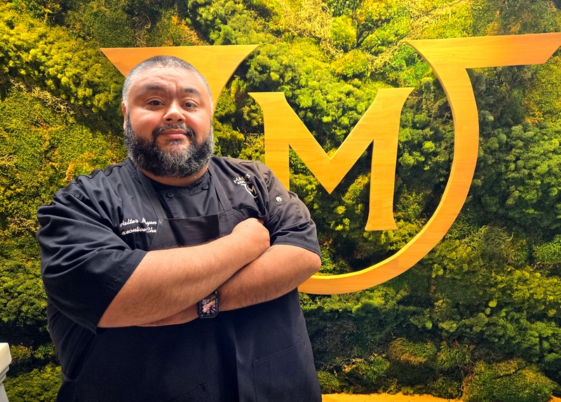 Walter Mayen, executive chef at Mastro's Steakhouse, standing with his arms crossed in a black shirt in front of a large gold G surrounded with plants