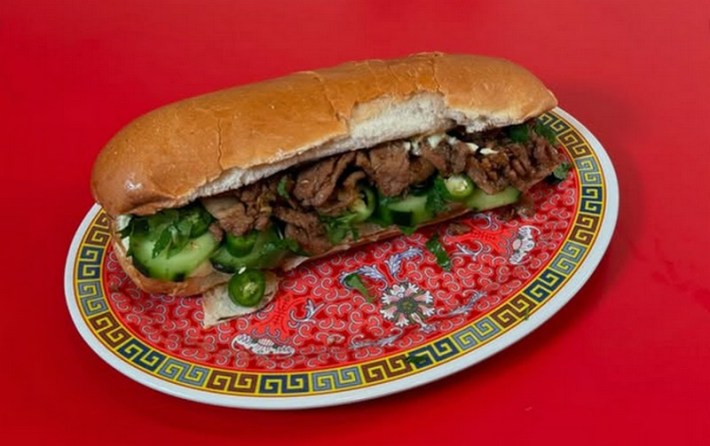 A beef banh mi on a roll with jalapenos and cucumbers sticking out of it, sitting on an ornate red plate with an Asian motif