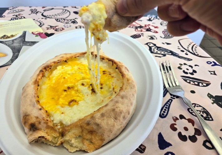 Cheese pulling from an Adjaruli khachapuri at Saqartvelo, next to a fork lying on the table, which is printed with a bird, flowers, and bean pod motifs.