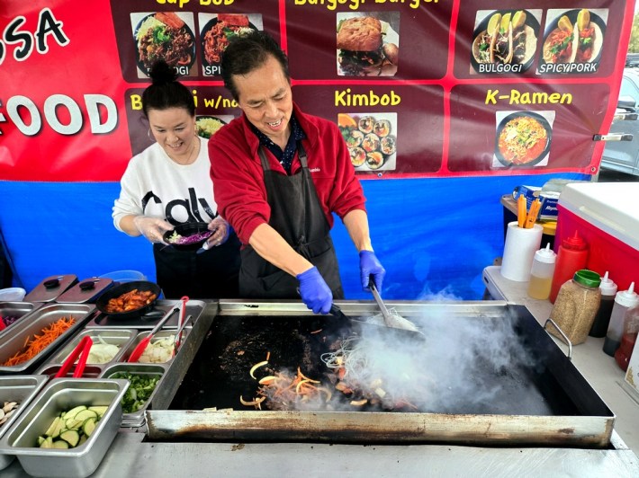 Assa's Savannah and Ken Lee, cooking side by side, and smiling, over a hot, smoking griddle.