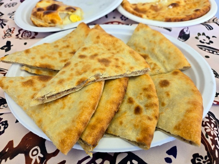 8 pinto bean-and-black pepper-filled lobiani flatbread stacked in a plate, with two plates of khachapuri sitting behind it.