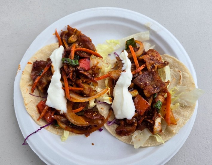 Two tacos with spicy pork, carrots, and a yogurt sauce on a white paper plate.