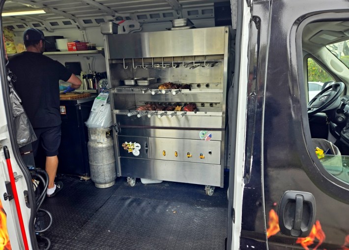 Looking into the side of a black van to see a stainless steel infrared rotisserie grill, with skewered meats. A man in shorts and a ballcap has his back to the camera on the left.