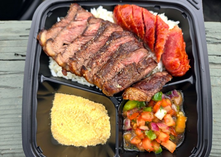 A black plastic tray of picanha steak and sausage over white rice, with sides of farofa (cassava flour) and molho a campanha.