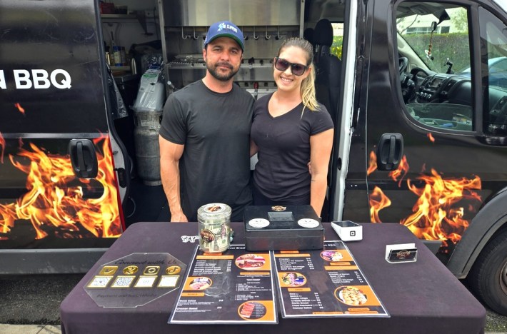 A man and a woman in black t-shirts stand in front of their van, which doubles as a Brazilian kitchen, with photos of flames depicted on its side.