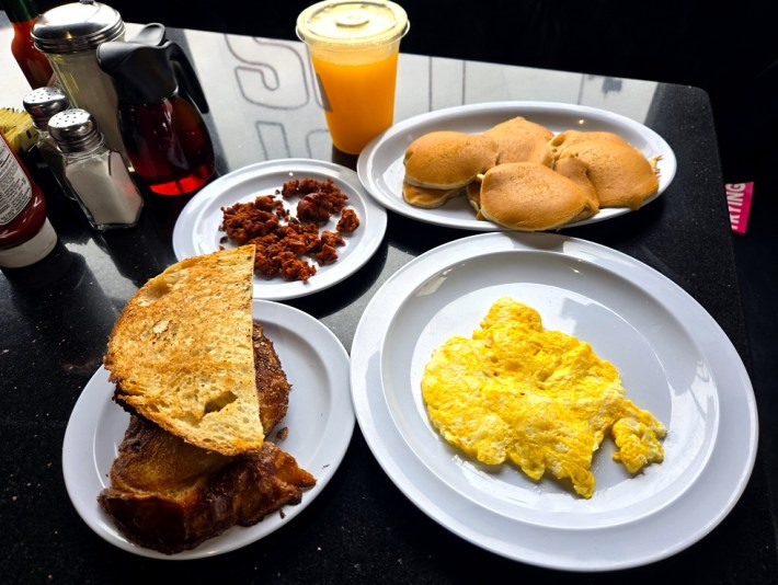 Garlic bread, chorizo, pancakes, and scrambled eggs on a black tabletop, next to a plastic cup of orange juice, salt, sugar, and pepper shakers, and a pitcher of maple syrup, at East Los Pantry