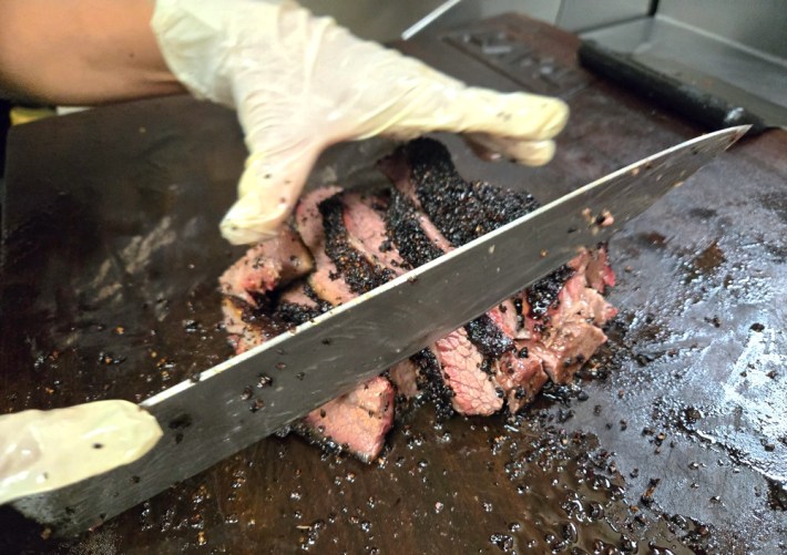 A large knife held by a gloved hand cuts into some sliced brisket