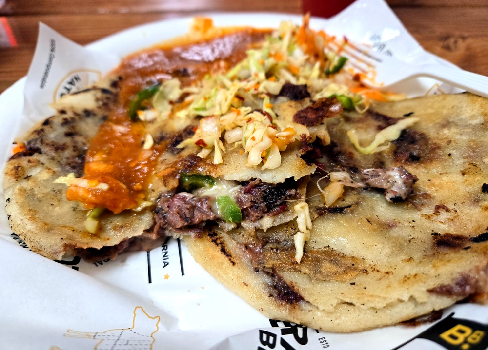 A pupusa cut in half showing brisket and red beans, with curtido and salsa all over the top.