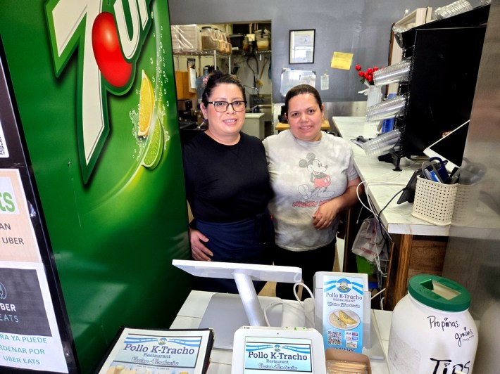 Carla Ramirez in glasses and a black shirt, with chef Sarita in a Mickey Mouse t-shirt, behind the counter at K-Tracho restaurant, beside a 7-11-branded fridge.