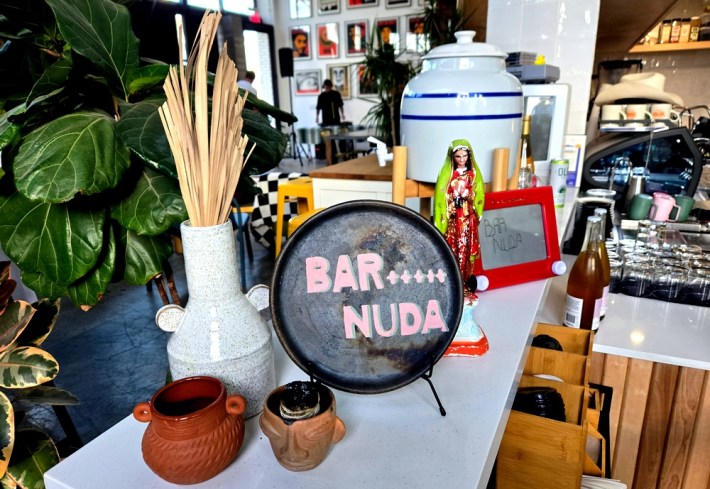 A countertop with a Virgen de Guadalupe statue, a vase full of dried palm, a plate that says Bar Nuda, and a ceramic water coooler and pots.