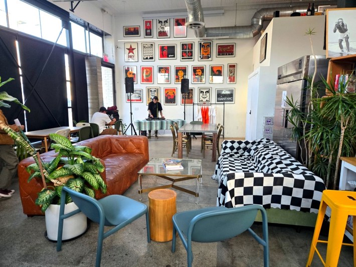 A large room with a checkered couch and leather couch, a DJ in front of numerous Shepard Fairey prints, and house plants