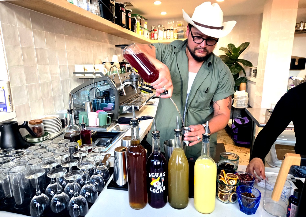 A bartender in a cowboy hat pours a red liquid from a bottle into a glass. In front of him are bottles with liquids in various colors.