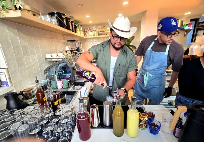 A man in a cowboy hat pours drinks next to a man in a Dodgers hat, looking at a coffee machine.