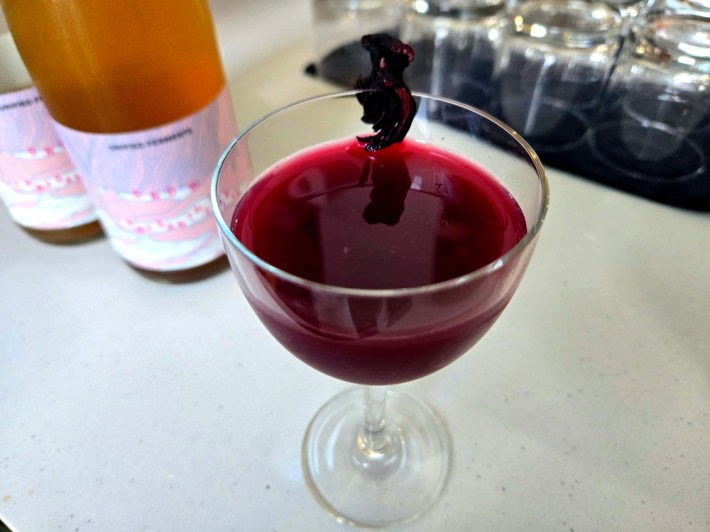 A deep red drink in a Nick and Nora glass garnished with dried hibiscus, in front of two bottles of an orange N/A wine.