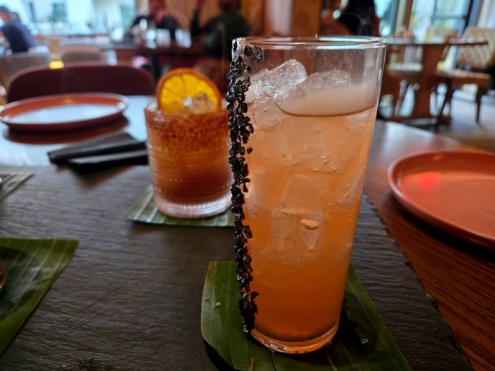 A light red cocktail with black salt vertically stuck to its side, and an orange slice-garnished drink behind it on the table