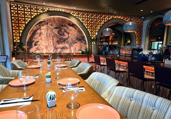 A long table leading up to an arcing mural of a guitar player in front of a church and next to a woman, next to a bar