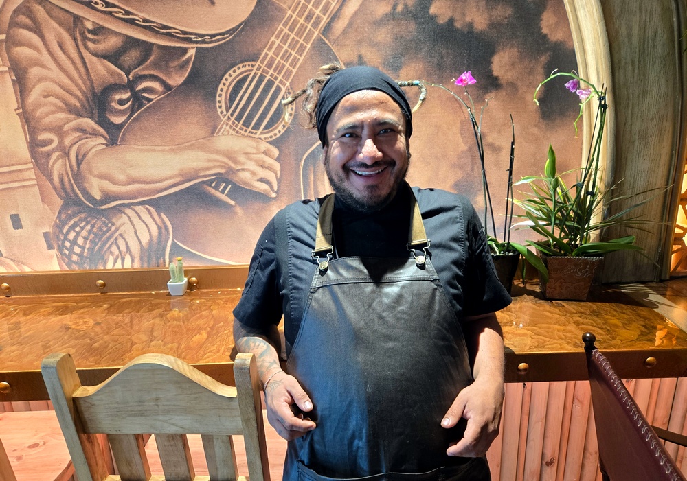 A chef with his dreadlocks tied behind in a headband stands with his elbows against a chair in front of a mural of a guitar player, smiling.