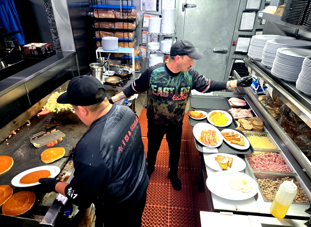 Original Pantry veterans Alex Ortiz and a cook named Pablo prepare breakfast at East Los Tacos., with Alex stretching across the kitchen to read an order as Pablo cooks pancakes on a griddle.